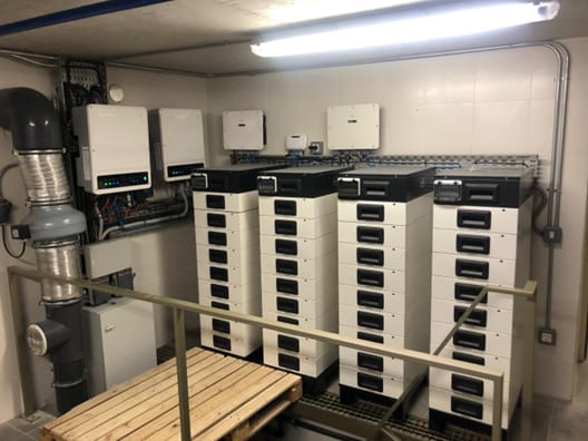 Plant room with four wall-mounted hybrid inverters above four floor-standing stacks of battery modules