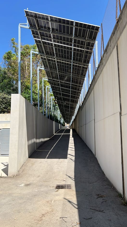 Solar canopy on steel supports running the length of a walled access road, seen from below against a clear sky